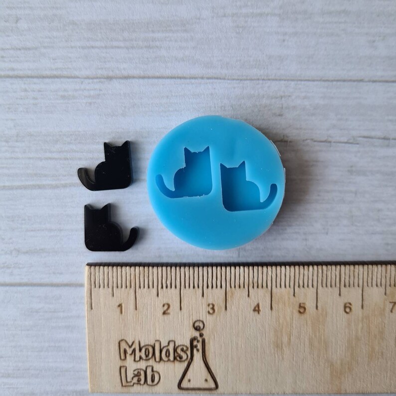 May include: A light blue silicone mold with two cat silhouettes, next to two black cat-shaped charms. A wooden ruler with inch markings is in the foreground. The text "Molds Lab" is visible.