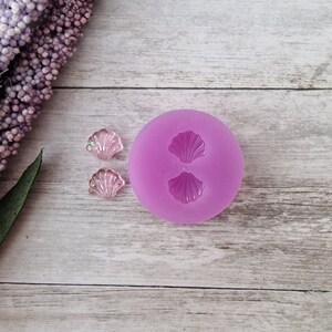May include: A lilac-colored silicone mold with two seashell impressions, next to two small, translucent seashell-shaped resin pieces with glitter. The items are on a light wood-grain surface, with a sprig of purple flowers.