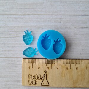 May include: A blue silicone mould featuring two strawberry shapes, with two translucent blue strawberry resin pieces. A wooden ruler with centimetre markings and the text "Molds Lab" is also present.