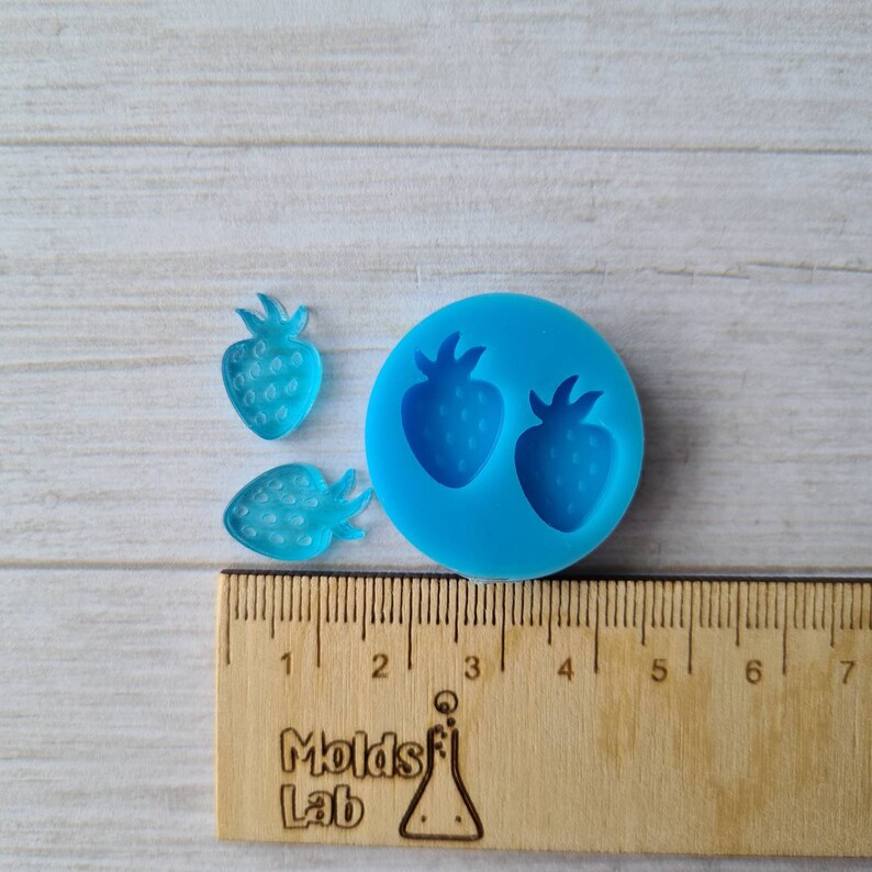 May include: A blue silicone mould featuring two strawberry shapes, with two translucent blue strawberry resin pieces. A wooden ruler with centimetre markings and the text "Molds Lab" is also present.