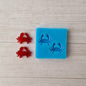 May include: Two red glitter crab-shaped embellishments next to a blue silicone mold with two crab designs. The mold is square-shaped. The background is a light wood grain.