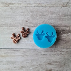 May include: Two rose gold glitter reindeer head earrings beside a blue silicone mould with two reindeer head impressions. The earrings are on a light wood grain surface.
