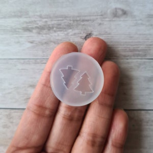 May include: A clear, circular silicone mould featuring two Christmas tree designs. One tree has a traditional shape, while the other has a more abstract, geometric form. The mould is held in a hand against a light wood grain background.