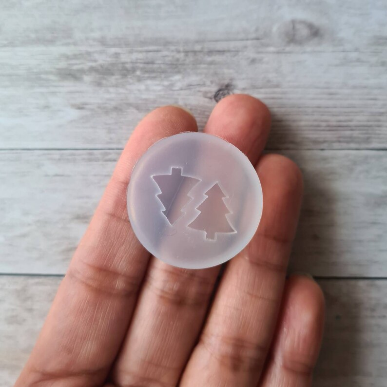 May include: A clear, circular silicone mould featuring two Christmas tree designs. One tree has a traditional shape, while the other has a more abstract, geometric form. The mould is held in a hand against a light wood grain background.