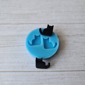May include: A light blue silicone mold with two cat silhouettes and two small black cat-shaped embellishments. The mold is on a light wood-grain surface. The cats are in a sitting position.