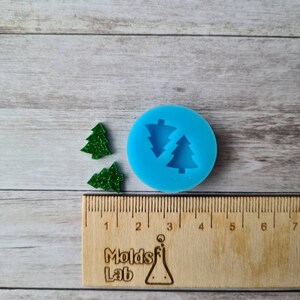May include: A blue silicone mould featuring two Christmas tree shapes, alongside two green glitter Christmas tree ornaments. A wooden ruler with the text "Molds Lab" is at the bottom. The background is a light wood grain.