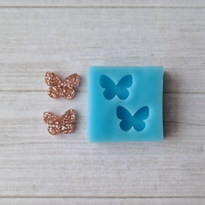 May include: A light blue silicone mould featuring two butterfly impressions, alongside two rose gold glitter butterfly embellishments. The butterflies are covered in glitter, and the mould is square.