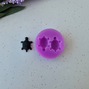 May include: A black turtle-shaped charm next to a purple silicone mould with two turtle impressions. The charm has a textured surface. The mould is round and has a smooth surface. The background is a speckled, light-coloured surface.