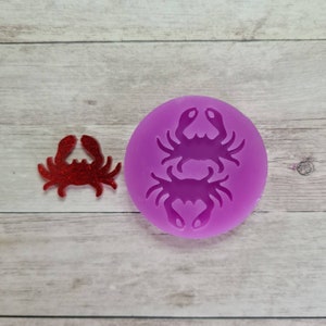 Puede incluir: Un cangrejo rojo con purpurina y un molde de silicona morado con un diseño de cangrejo. El cangrejo es un pequeño objeto decorativo. El molde es redondo y presenta una silueta detallada de cangrejo, ideal para proyectos de manualidades.