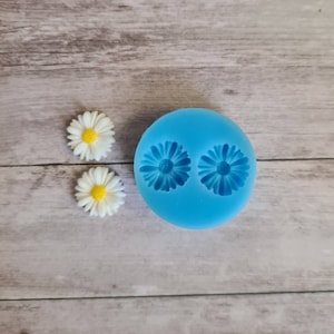 May include: A blue silicone mould with two daisy flower impressions, next to two finished daisy flowers. The flowers have white petals and yellow centres. The mould and flowers are on a light wood grain surface.