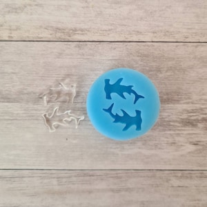 Könnte beinhalten: Eine hellblaue Silikonform mit zwei Hammerhai-Formen. Zwei transparente, haiförmige Objekte befinden sich neben der Form. Die Objekte sind wahrscheinlich das Ergebnis der Verwendung der Form. Der Hintergrund ist eine helle Holzmaserung.