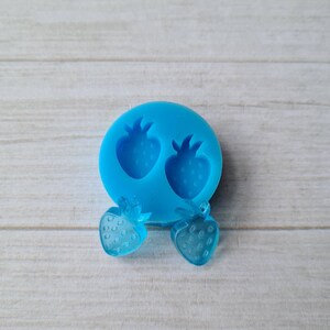 May include: A light blue silicone mould with two strawberry impressions, alongside two transparent strawberry-shaped resin charms. The charms and mould are on a light wood-grain surface. The strawberry charms have visible details.