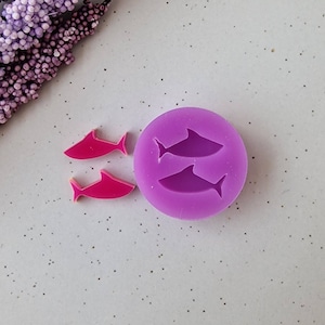 Puede incluir: Primer plano de un molde de silicona morado con dos siluetas de tiburones y dos piezas rosas con forma de tiburón. El molde es redondo y los tiburones miran en la misma dirección. El fondo es una superficie clara y moteada.