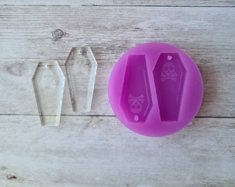 May include: Clear coffin-shaped pendants with skull and crossbones design, beside a purple silicone mould with coffin cavities. The pendants are made of clear resin. The mould has two coffin shapes with skull and crossbones.