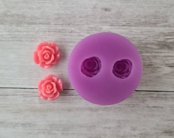 May include: A purple silicone mold with two rose-shaped cavities, next to two pink rose-shaped resin castings. The mold and castings are on a light wood-grain surface. The rose castings are a vibrant pink color.