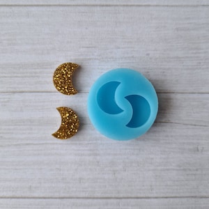 May include: A light blue silicone mould featuring two crescent moon shapes, alongside two gold glitter crescent moon charms. The mould and charms are on a white wooden surface. The charms are approximately 2.5 cm in length.