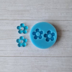 May include: A light blue silicone mould featuring two flower-shaped cavities, with two transparent blue flower-shaped resin pieces. The circular mould and resin pieces have a central hole. The items are on a light wood grain surface.
