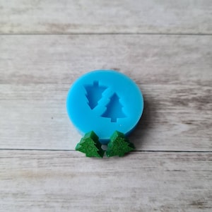 May include: A light blue silicone mould featuring two Christmas tree shapes. Two small, green, glittery Christmas tree-shaped resin pieces are beside the mould. The background is a light wood grain.