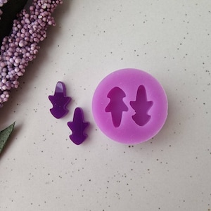 Puede incluir: Primer plano de un molde de silicona morado con dos cavidades, cada una con forma de árbol o hoja estilizada. Dos pequeñas piezas de resina morada terminadas están junto al molde. El fondo es de color neutro.