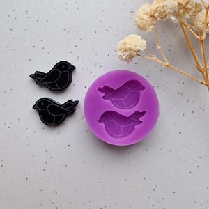 May include: Two black bird-shaped embellishments and a purple silicone mold with two bird impressions. The embellishments are glossy and have a simple, stylized design. The mold is round and features the same bird shape.