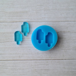 Pode incluir: Um molde de silicone azul com duas formas de picolé e duas peças de acrílico azul transparente em forma de picolé. Os itens estão em uma superfície de grão de madeira clara. O molde é redondo e tem uma textura lisa.