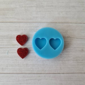 May include: A light blue silicone mould with two heart-shaped cavities, beside two small, red glitter heart-shaped embellishments. The items are on a light wood-grain surface. The mould is round and the hearts are solid.