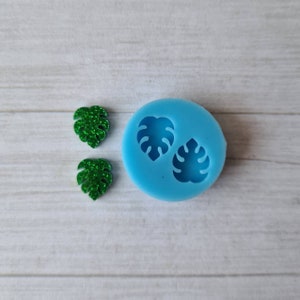 May include: A light blue silicone mould with two leaf-shaped cavities, next to two green glitter leaf-shaped embellishments. The mould and embellishments are on a white wooden surface. The leaves are a tropical theme.