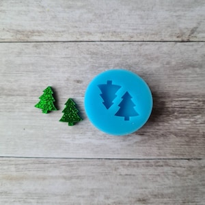 May include: A blue silicone mould featuring two Christmas tree shapes, alongside two small, glittery green Christmas tree ornaments. The ornaments are a vibrant green with a sparkly texture. The mould is round and has a smooth surface.