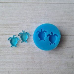 May include: Two small, transparent blue turtle-shaped charms beside a round, bright blue silicone mould with two turtle impressions. The items are on a light wood-grain surface. The charms are likely for jewellery or craft projects.