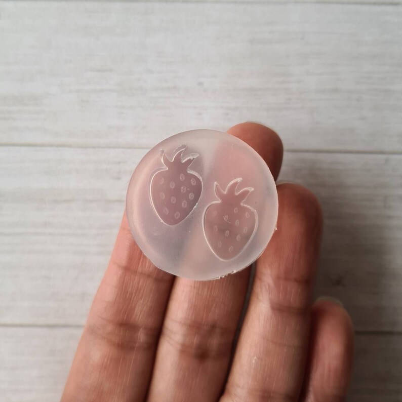 May include: A clear, circular mould featuring two strawberry designs. The strawberries are outlined with detailed stems and seeds. This mould is likely used for crafting or creating decorative items.