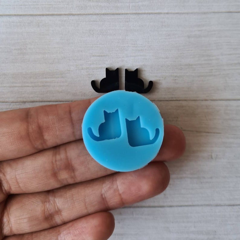 May include: A light blue silicone mold with two cat silhouettes, along with two black cat-shaped cutouts. The items are displayed on a light wood-grain surface. The mold is round and the cat shapes are facing each other.