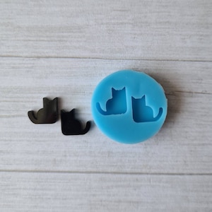 May include: A light blue silicone mold with two cat silhouettes, next to two black cat-shaped cutouts. The mold is round, and the cats are sitting with their tails curled. The cutouts are solid black and have a glossy finish.