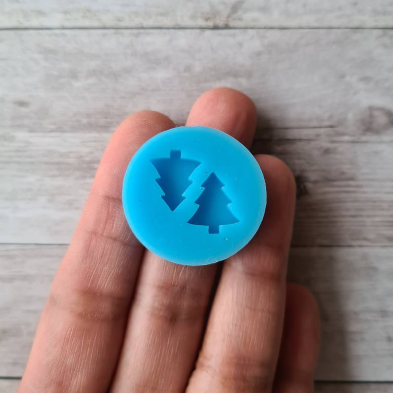 May include: A circular, vibrant blue silicone mould displaying two Christmas tree designs. The mould is held in a hand, with a light wood grain background. The trees are detailed with triangular shapes, ideal for crafting.