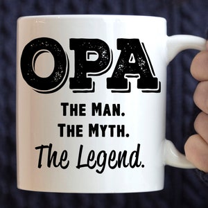 May include: White ceramic mug with the word "OPA" in large, distressed black letters. Below, the text reads "THE MAN. THE MYTH. The Legend." The mug has a curved handle and is held by a person.