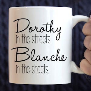 May include: White ceramic coffee mug with black text that reads "Dorothy in the streets. Blanche in the sheets."