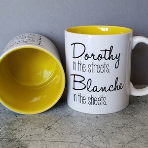 May include: White ceramic mug with a yellow interior and black text that reads "Dorothy in the streets. Blanche in the sheets."