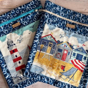 May include: Two rectangular fabric pouches with a dark blue bubble pattern border. One pouch features a red and white lighthouse, the other displays three beach huts, a deck chair, and a red umbrella. Both have zips and a wooden label reading Made by J Green Lane Crafts.