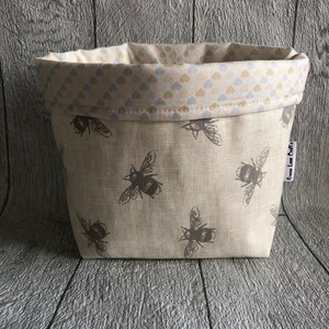 Peut inclure: Un petit panier en tissu beige orn&eacute; d'un motif r&eacute;p&eacute;t&eacute; d'abeilles grises. La doublure int&eacute;rieure est de couleur cr&egrave;me avec un motif de petits c&oelig;urs bleus et beiges. Le panier porte une &eacute;tiquette "Gwen Love Crafts".