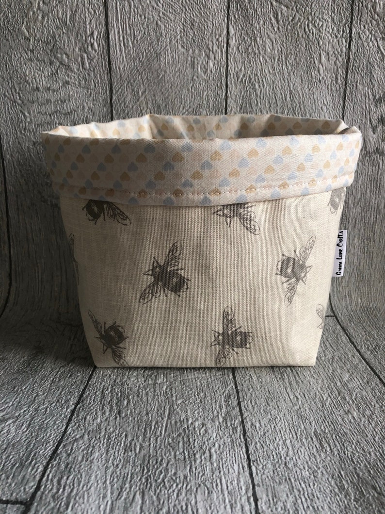 Peut inclure: Un petit panier en tissu beige orn&eacute; d'un motif r&eacute;p&eacute;t&eacute; d'abeilles grises. La doublure int&eacute;rieure est de couleur cr&egrave;me avec un motif de petits c&oelig;urs bleus et beiges. Le panier porte une &eacute;tiquette "Gwen Love Crafts".
