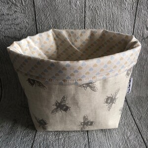 Peut inclure: Un panier en tissu beige orn&eacute; de motifs d'abeilles grises. L'int&eacute;rieur est doubl&eacute; d'un motif de petits c&oelig;urs bleus et beiges. Le panier est de forme rectangulaire avec un bord sup&eacute;rieur repli&eacute;.