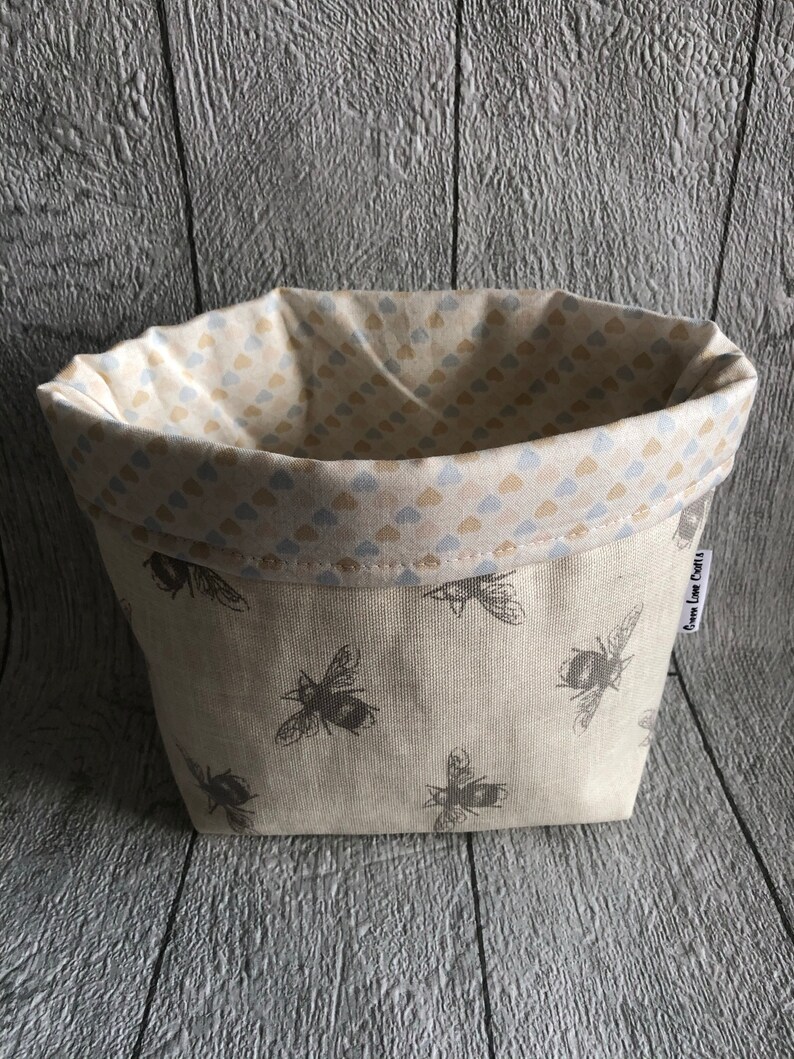 Peut inclure: Un panier en tissu beige orn&eacute; de motifs d'abeilles grises. L'int&eacute;rieur est doubl&eacute; d'un motif de petits c&oelig;urs bleus et beiges. Le panier est de forme rectangulaire avec un bord sup&eacute;rieur repli&eacute;.