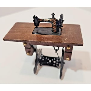 Op de afbeelding: Een miniatuurnaaimachine met een zwarte metalen behuizing met gedetailleerde gouden en rode bloemmotieven, gemonteerd op een rechthoekig bruin houten tafelblad ondersteund door een zwart metalen frame met een decoratieve pedaalvoet en gouden beslag.