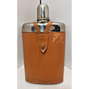 Può includere: Una fiaschetta in acciaio inossidabile con custodia in pelle marrone. La fiaschetta ha un tappo argentato e un cinturino in pelle con chiusura a bottone. La custodia in pelle mostra usura.