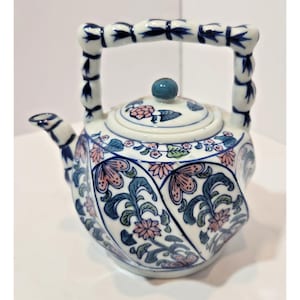 Puede incluir: Una tetera de cerámica blanca con un patrón floral azul y rosa con hojas verdes. La tetera tiene un asa estilo bambú y una tapa a juego con un pequeño pomo azul. El pico es curvo y está decorado con detalles azules a juego con el asa.