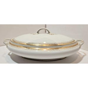 Puede incluir: Fuente ovalada de porcelana blanca con tapa y asas. La fuente presenta un ribete dorado a lo largo del borde y la tapa, con un asa dorada en la parte superior. La fuente está diseñada para servir alimentos.