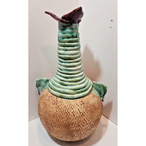 May include: A handcrafted ceramic vase with a textured, speckled tan base and a ribbed, turquoise neck. The vase features a dark burgundy, petal-like accent at the top and turquoise handles. This unique piece is a decorative art object.