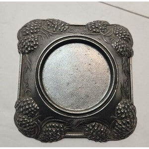 May include: Square, silver-toned metal coaster with a central circular depression. The coaster's corners feature detailed relief carvings of pinecones and foliage, adding a decorative touch. The metal has a slightly aged appearance.