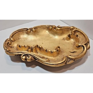 May include: Ornate gold-colored ashtray with a decorative, scrolled edge. The ashtray has a shallow, oval shape with a textured surface and several built-in cigarette rests. The overall design is vintage and elegant.