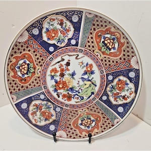 Puede incluir: Un plato decorativo redondo de cerámica que presenta un medallón circular central con un pájaro posado en una rama entre flores naranjas y amarillas. El borde está dividido en paneles alternos de patrones geométricos azul oscuro y naranja.