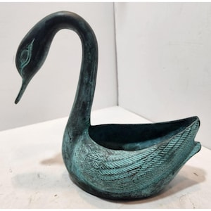 Op de afbeelding: Een teal-kleurige zwaanvormige decoratieve kom. De zwaan heeft een lange, gebogen nek en een gedetailleerd veermotief op zijn vleugels. De kom is gemaakt van donker metaal met een verweerde, verouderde uitstraling.